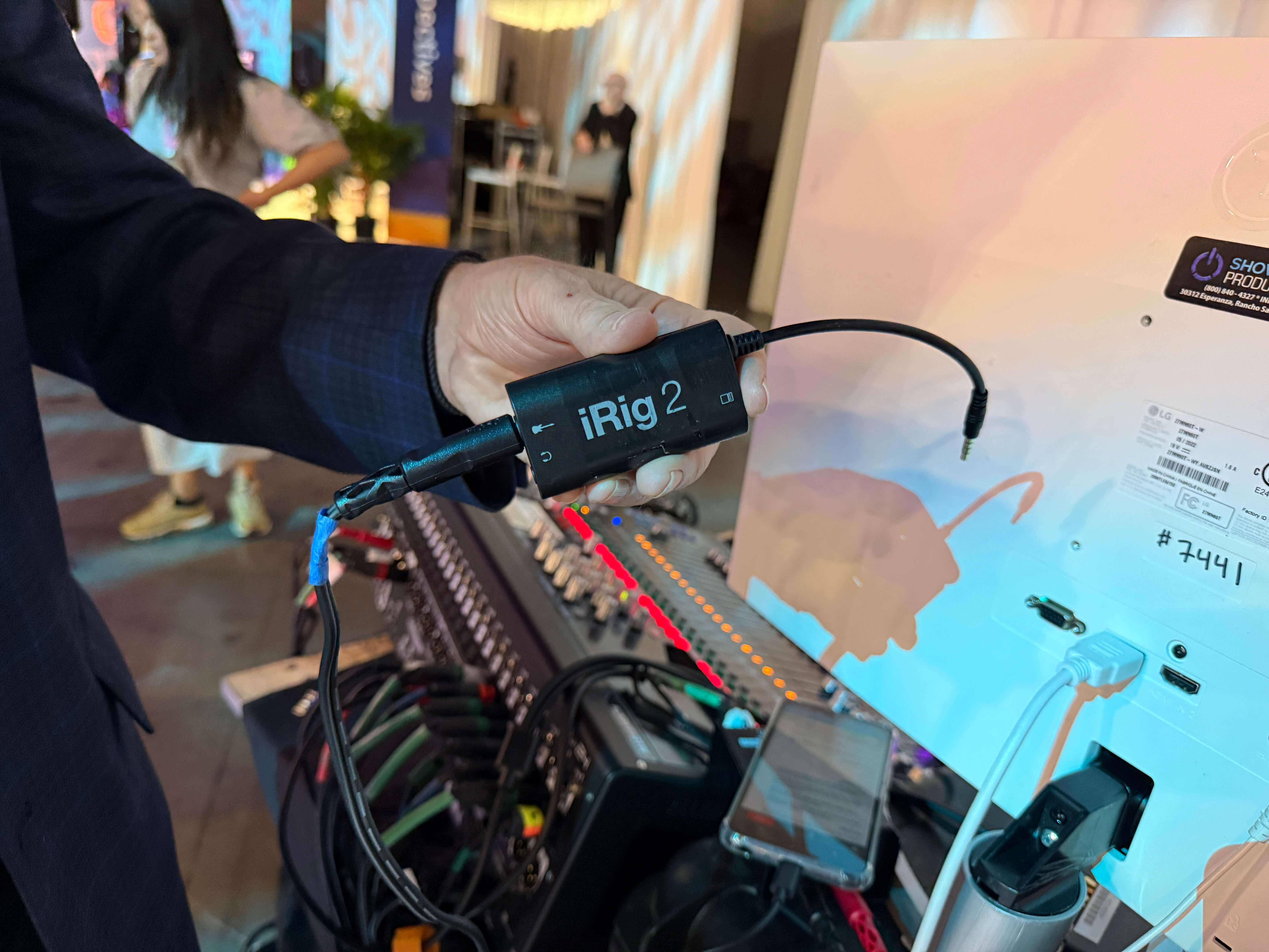 A person holds an iRig 2 audio converter device connected to an audio mixer with multiple cables during an event setup. The iRig is shown as part of a Wordly AV connection, linking the mixer to a smartphone for real-time audio input to the Wordly AI system.